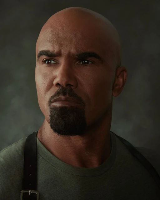 Close-up portrait of an adult male with a shaved head and goatee wearing a green shirt and black suspenders against a gray backdrop.